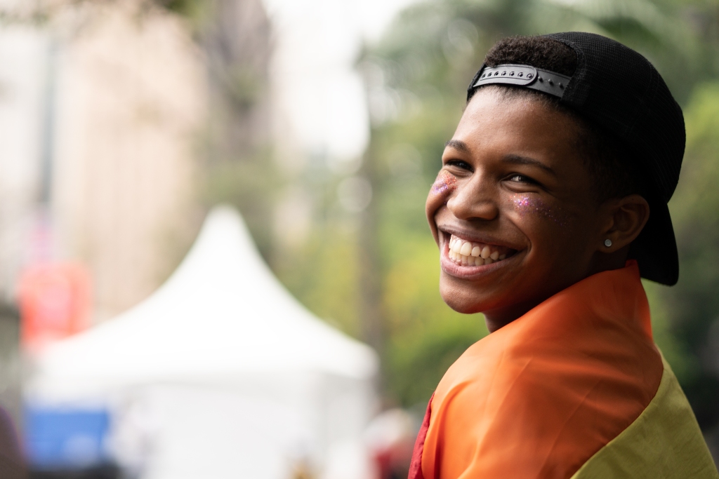 Photo of a Black boy smiling widely. He has purple glitter on his cheeks and a rainbow Gay Pride flag over his shoulders. The background is blurry, with trees and a white event tent.